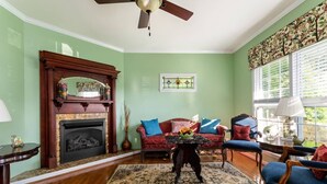 A living room with a fireplace, a ceiling fan, a large window with blinds, and a patterned rug.