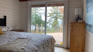 Une chambre à coucher comprenant un lit, une tête de lit en bois, une table de chevet, une lampe et une vue sur une forêt et un plan d’eau, visible à travers une porte-fenêtre coulissante.