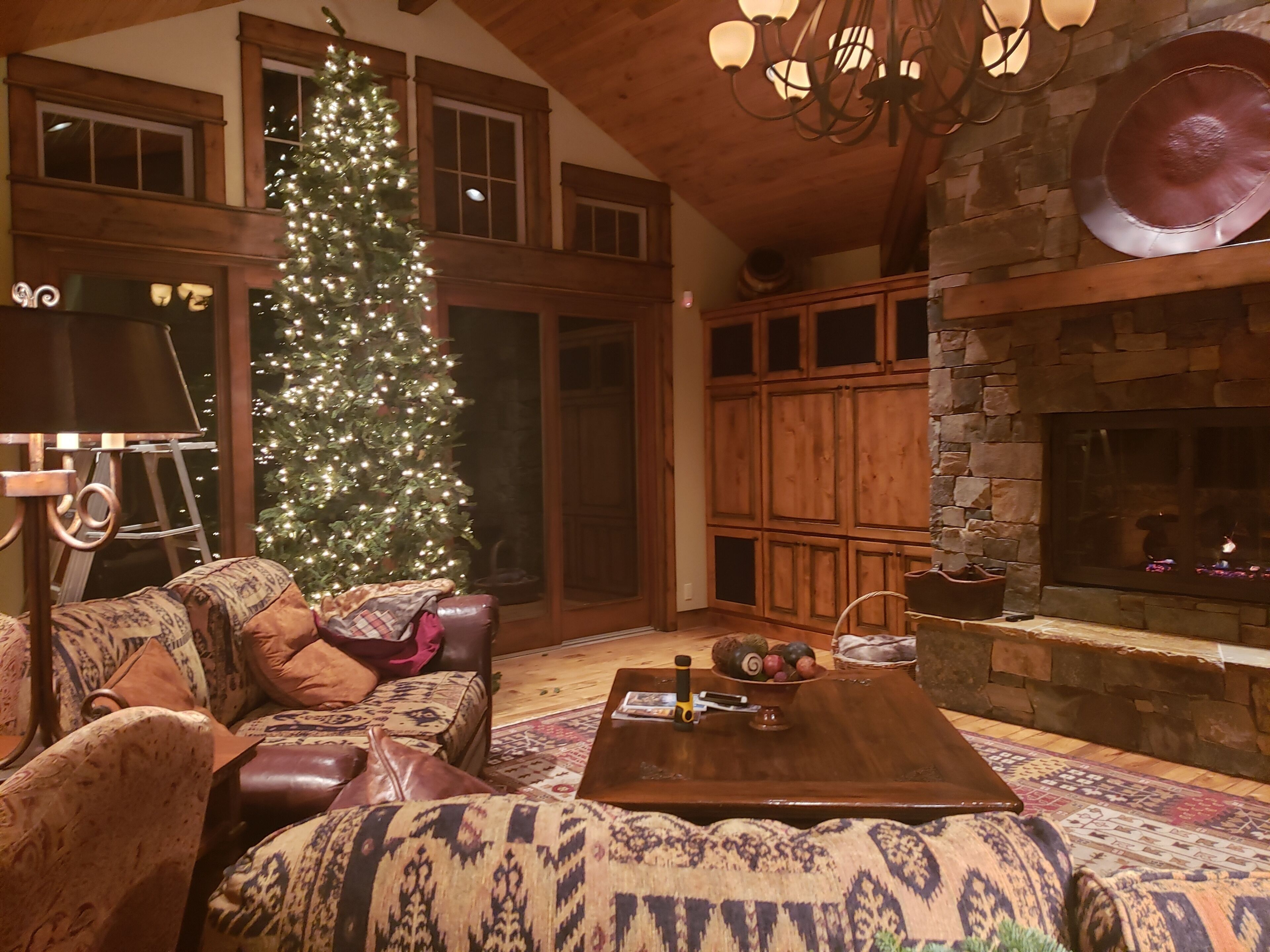 A cozy living room with a large Christmas tree, leather sofas, a wooden coffee table, and a stone fireplace.