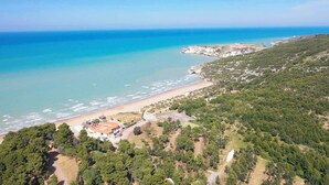 An aerial view of a coastal area with a beach, dense forest, and a small cluster of buildings with a red roof.