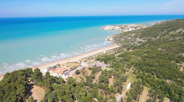 Vista aérea de una zona costera con playa, bosque denso y un pequeño conjunto de edificios con techo rojo.