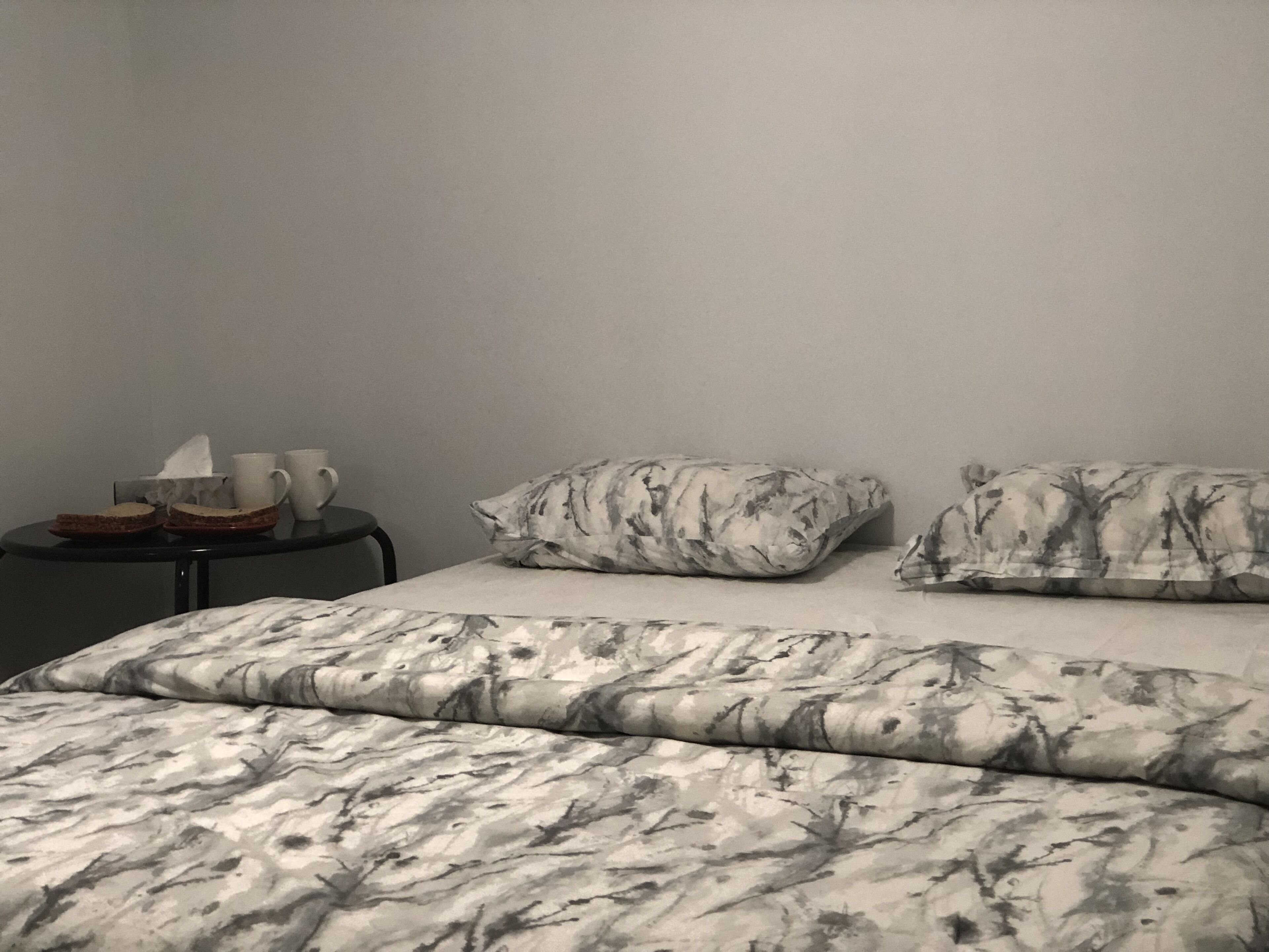 A hotel room with a bed featuring a marble pattern, two pillows with a similar design, a small round table with a tissue box, a cup, and a sandwich.