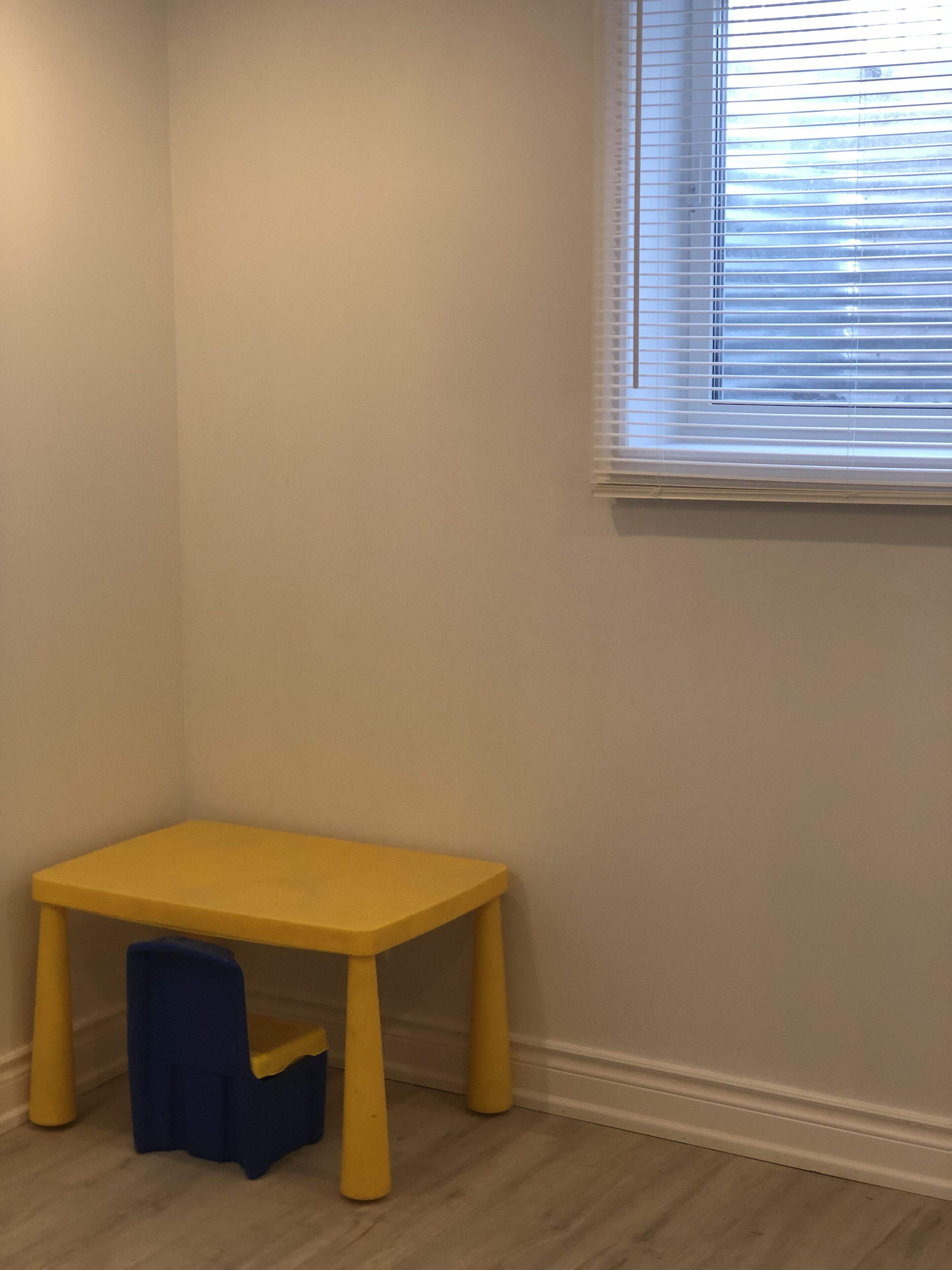 A corner room with a yellow table and a blue chair, a window with blinds, and a plain wall.