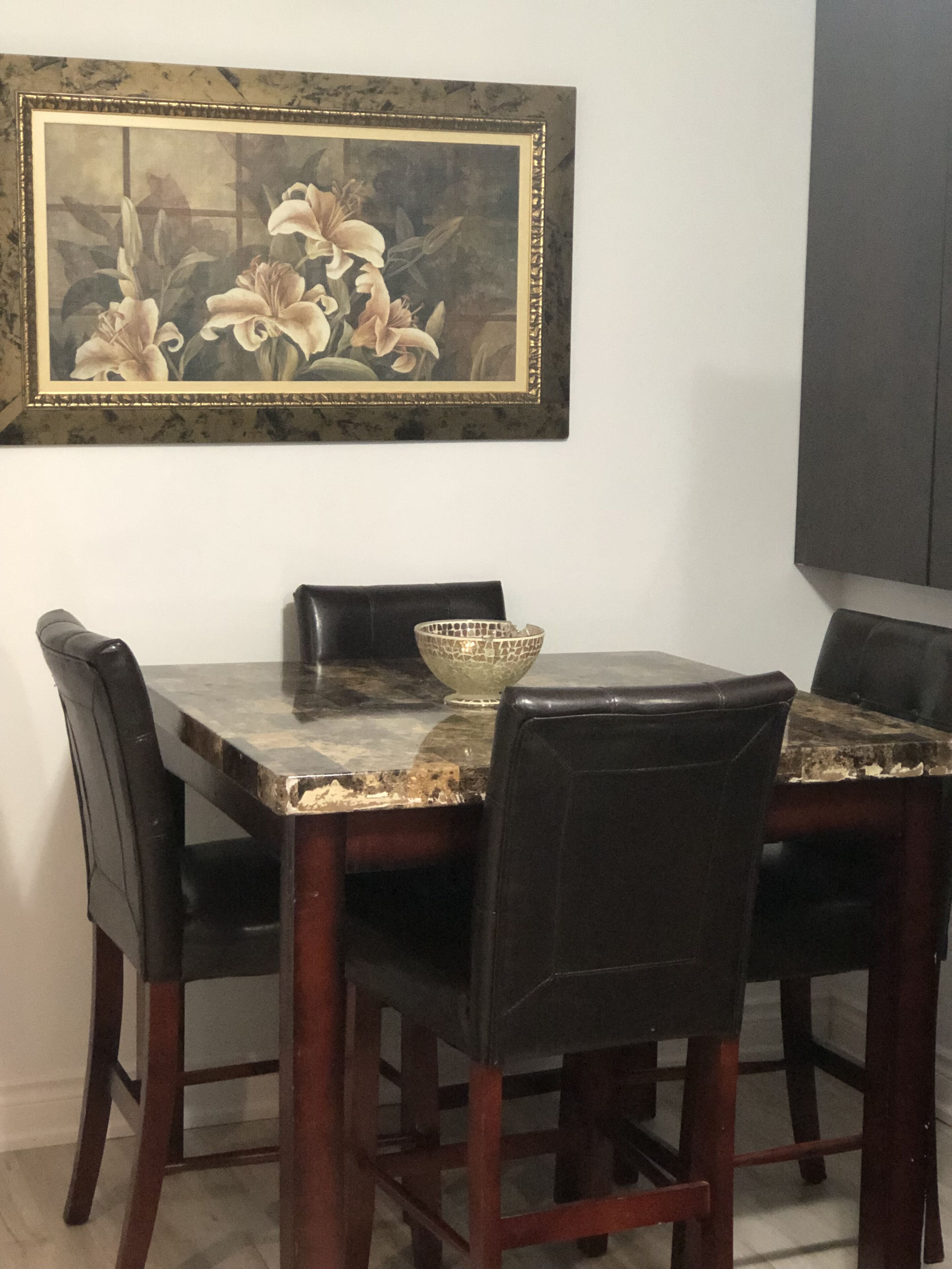 A dining table with a marble top, surrounded by four chairs. A framed painting of flowers hangs above the table.