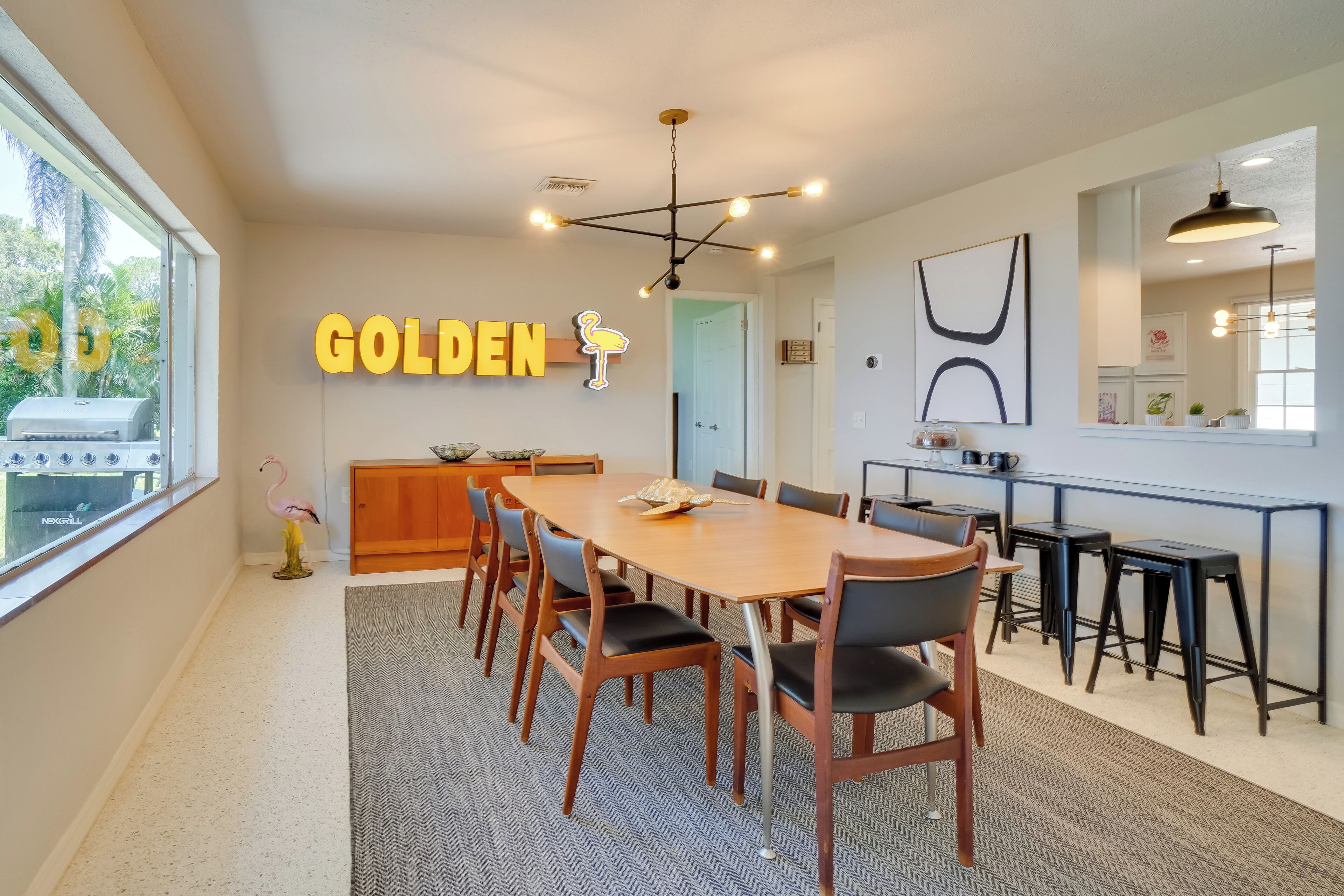 A modern dining area with a wooden table and chairs, a neon sign reading "GOLDEN," and a view of the outdoors through large windows.