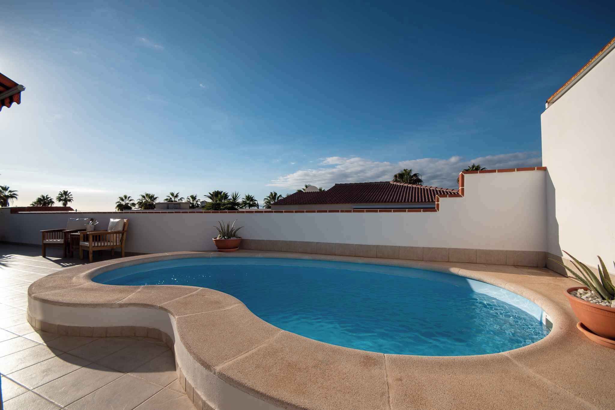 A rooftop swimming pool with a curved edge, surrounded by a white wall and a tiled floor. There are potted plants and a wooden table with chairs.