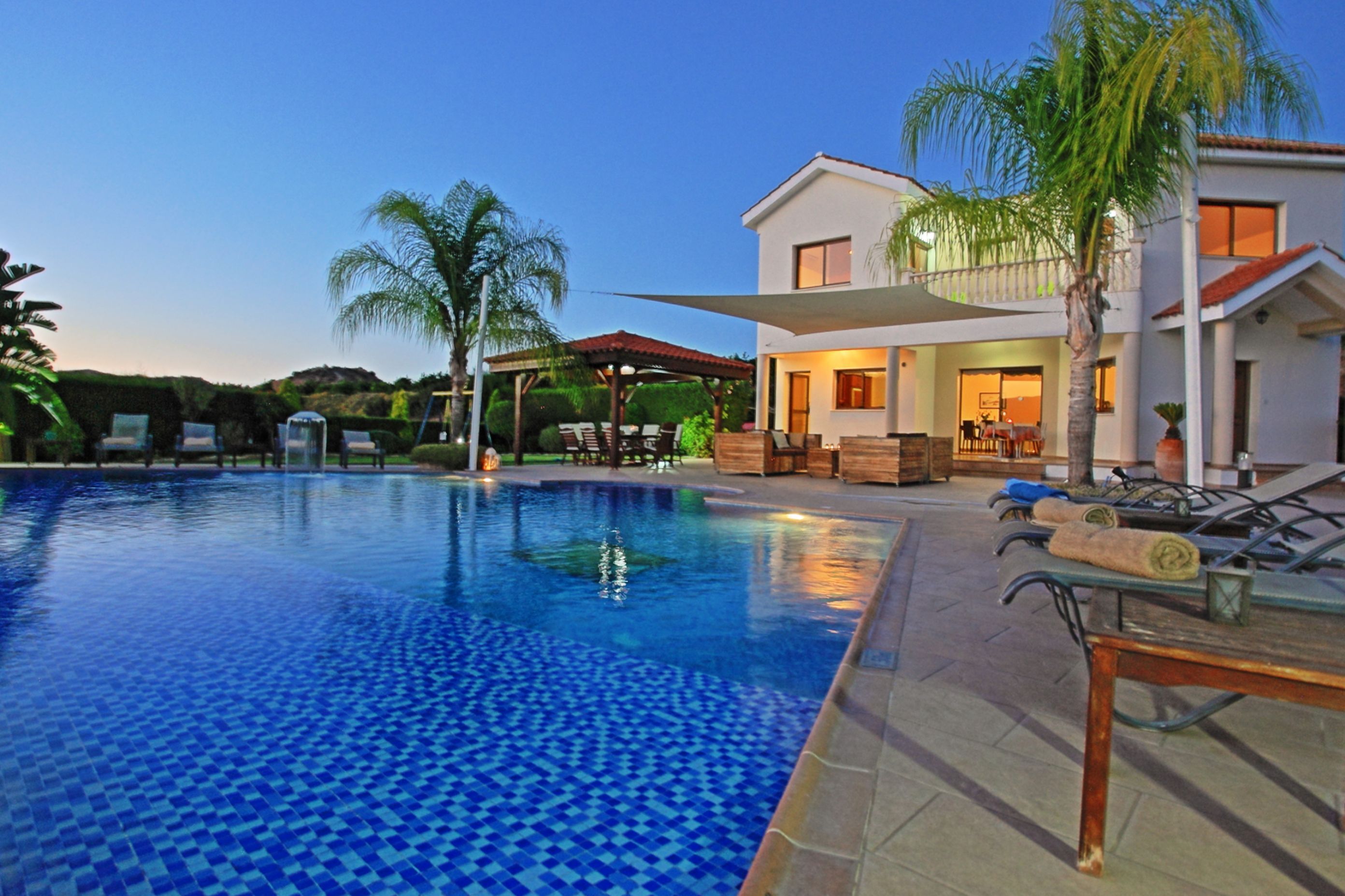 Ein Swimmingpool mit Liegestühlen und Sonnenschirmen, ein überdachter Bereich mit Sitzgelegenheiten und ein zweistöckiges Gebäude mit großen Fenstern.