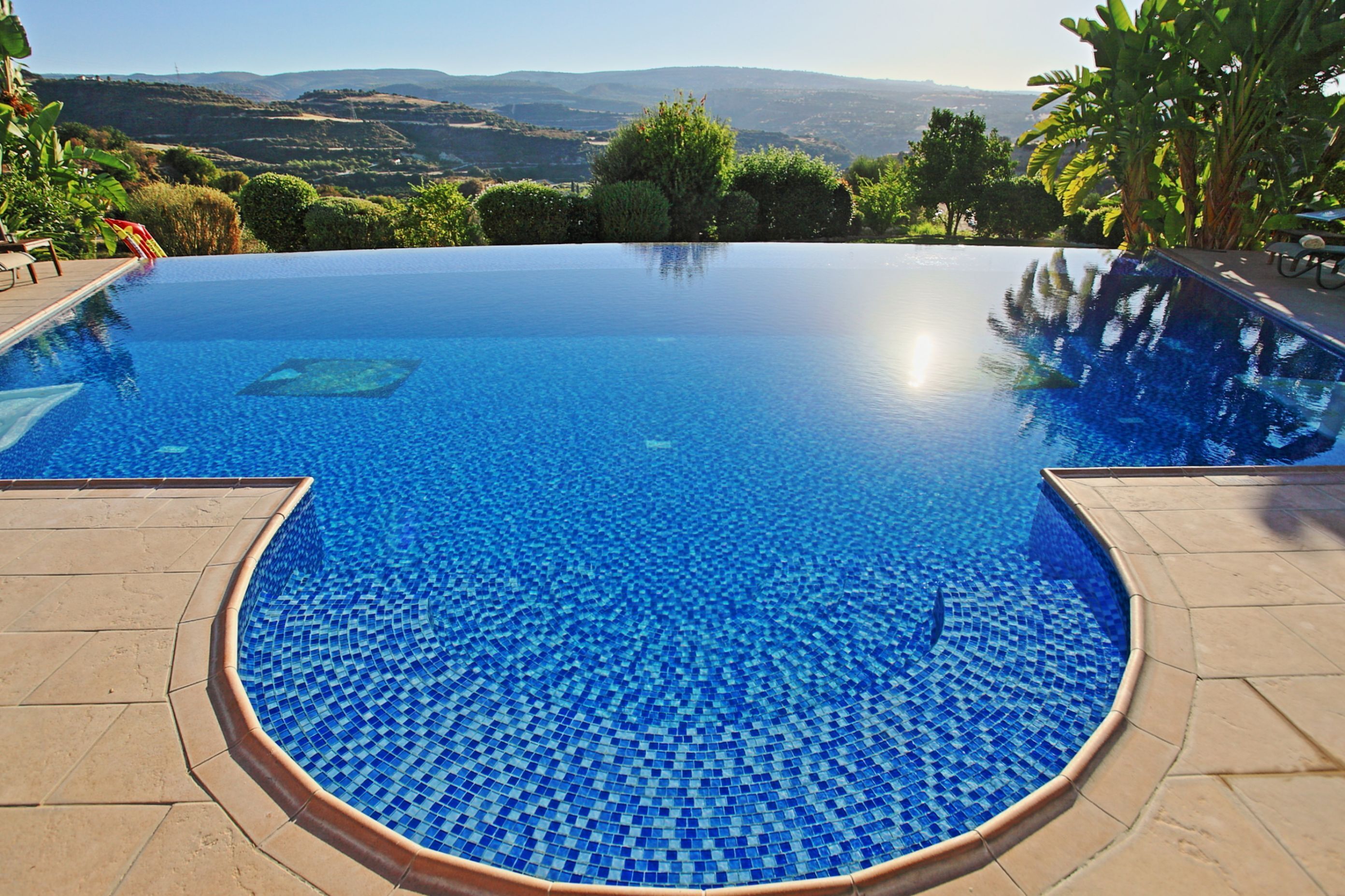 A clear view of a swimming pool with a tiled edge, surrounded by a paved deck, and overlooking a scenic landscape.