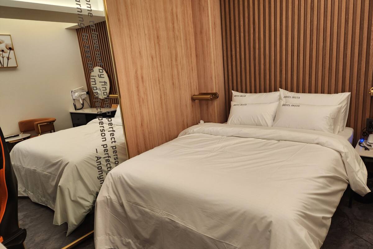 A hotel room with two beds, a wooden panel wall, a small desk with a lamp, and a chair.