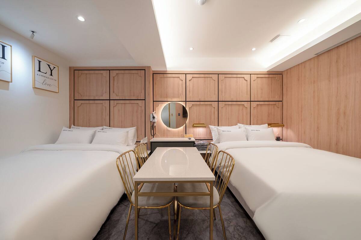 A hotel room with two beds, a dining table with chairs, a mirror, and wooden paneling on the walls.