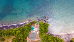 Vista aérea de uma propriedade costeira com piscina, área de varanda e vegetação exuberante.