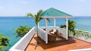 Um deck de madeira com um gazebo, corrimãos brancos e vista para o oceano.