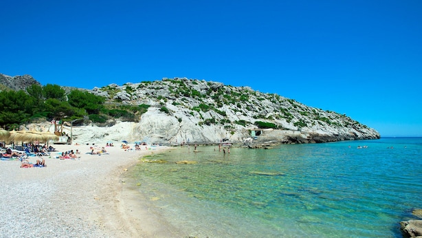 En strand med klart vand, folk der solbader, og en klippe i baggrunden.