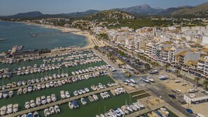 A marina with numerous boats docked, a beach area, and a coastal town with buildings and mountains in the background.