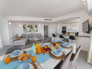 Een moderne woonkamer met een bank, een eethoek met een tafel gedekt voor het ontbijt, en een keuken met witte kasten.