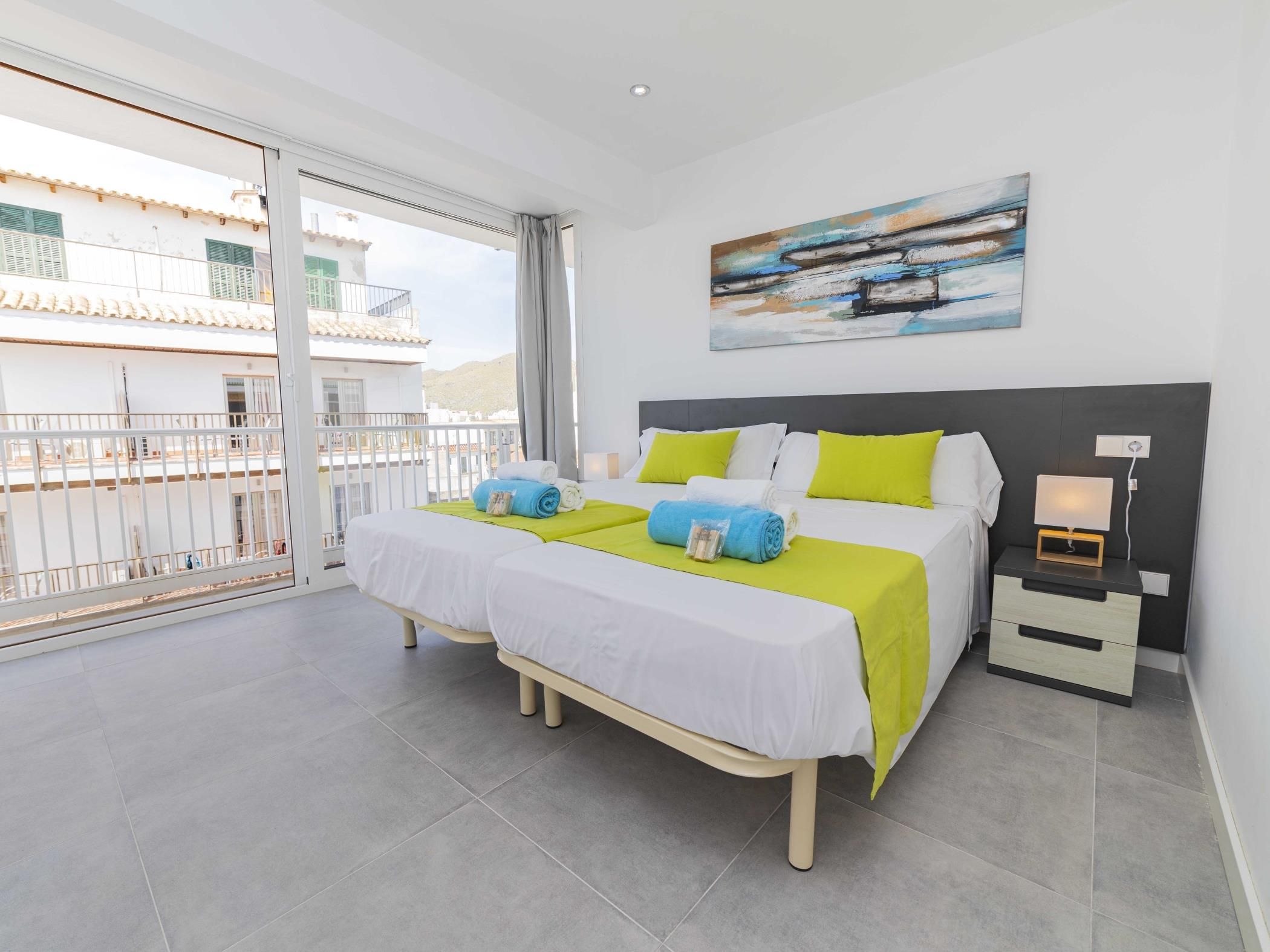 A hotel room with a balcony, a bed with two pillows, a bedside table with a lamp, and a painting on the wall.