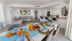 A modern dining area with a long table set for breakfast, a living room with a gray sofa, and a kitchen in the background.