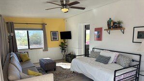 Ein modernes Wohnzimmer mit einer Couch, einem Hocker und einem Bett mit Kopfteil. Es gibt einen Deckenventilator, einen Fernseher und einen kleinen Tisch mit einem Buch.