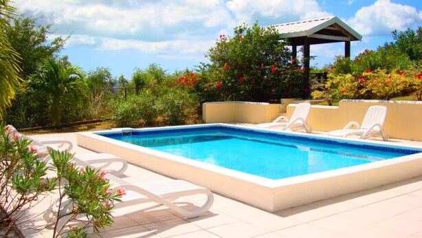 Une piscine rectangulaire dont les bords sont recouverts de carreaux, entourée d’une terrasse également carrelée. On peut y voir deux chaises longues blanches, et une structure couverte avec un toit est visible à l’arrière-plan.