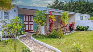 Un bâtiment blanc avec une porte rouge, entouré d’un jardin où poussent différentes espèces de plantes et bordé d’un chemin en pierre.
