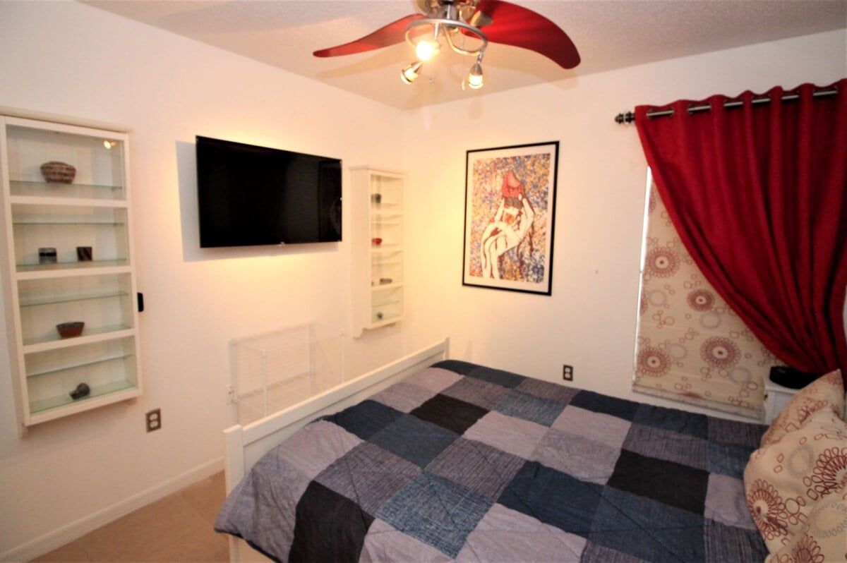 Quarto com cama, TV fixada na parede, ventilador de teto, cortina vermelha e prateleira com objetos decorativos.