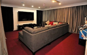 A living room with a large flat-screen TV mounted on the wall, a sectional sofa, a dark-colored entertainment unit, and a red carpet. - Red Corvette House with Theater Room - WR001 (Davenport)