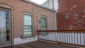 Una terraza de madera con barandilla metálica, un muro de ladrillo y un edificio con dos ventanas y una puerta.