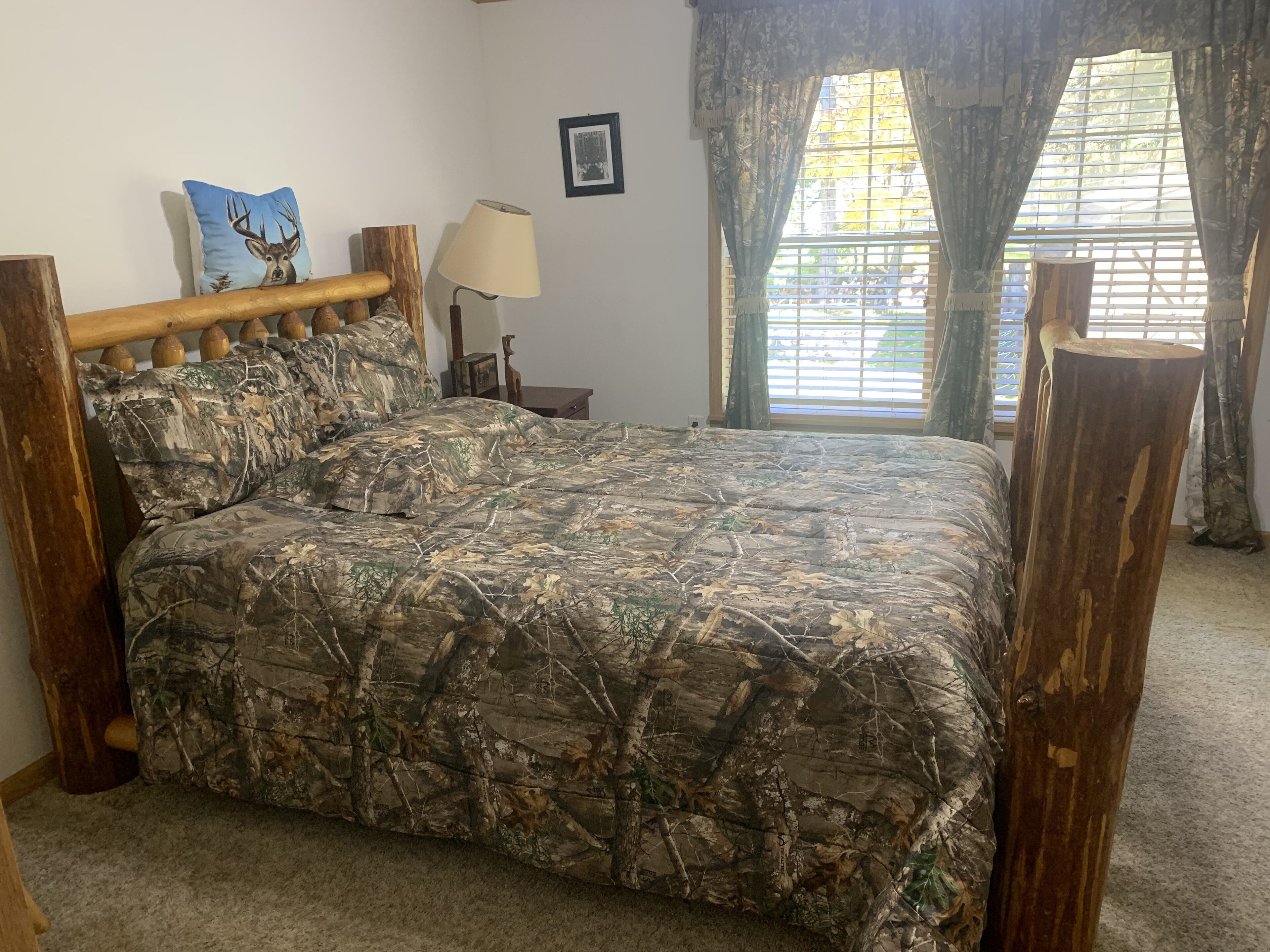 Un dormitorio con edredón estampado de camuflaje, cabecero de madera, una lámpara y una ventana con cortinas.