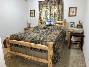 Een slaapkamer met een bed met camouflageprint, een nachtkastje met een lamp, een houten bureau en twee ingekaderde foto's aan de muur.