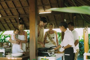 A group of people around a food preparation area with a rustic thatched roof. - The Kelong Trikora Resort (Bintan)
