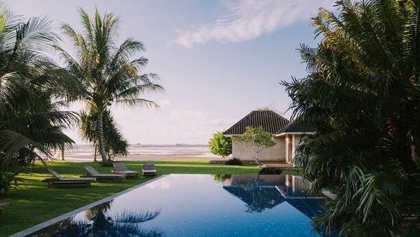 A swimming pool with lounge chairs and palm trees, a building with a sloped roof, and a beach in the background. - The Kelong Trikora Resort (Bintan)