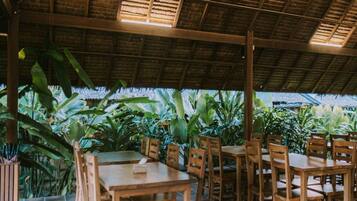 An outdoor dining area with wooden tables and chairs, surrounded by lush tropical plants.