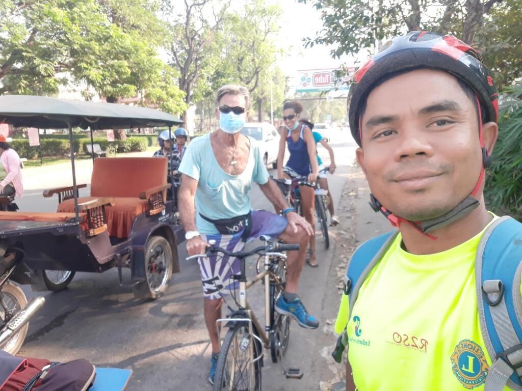 A group of cyclists on a street with a vintage car and a tuk-tuk.