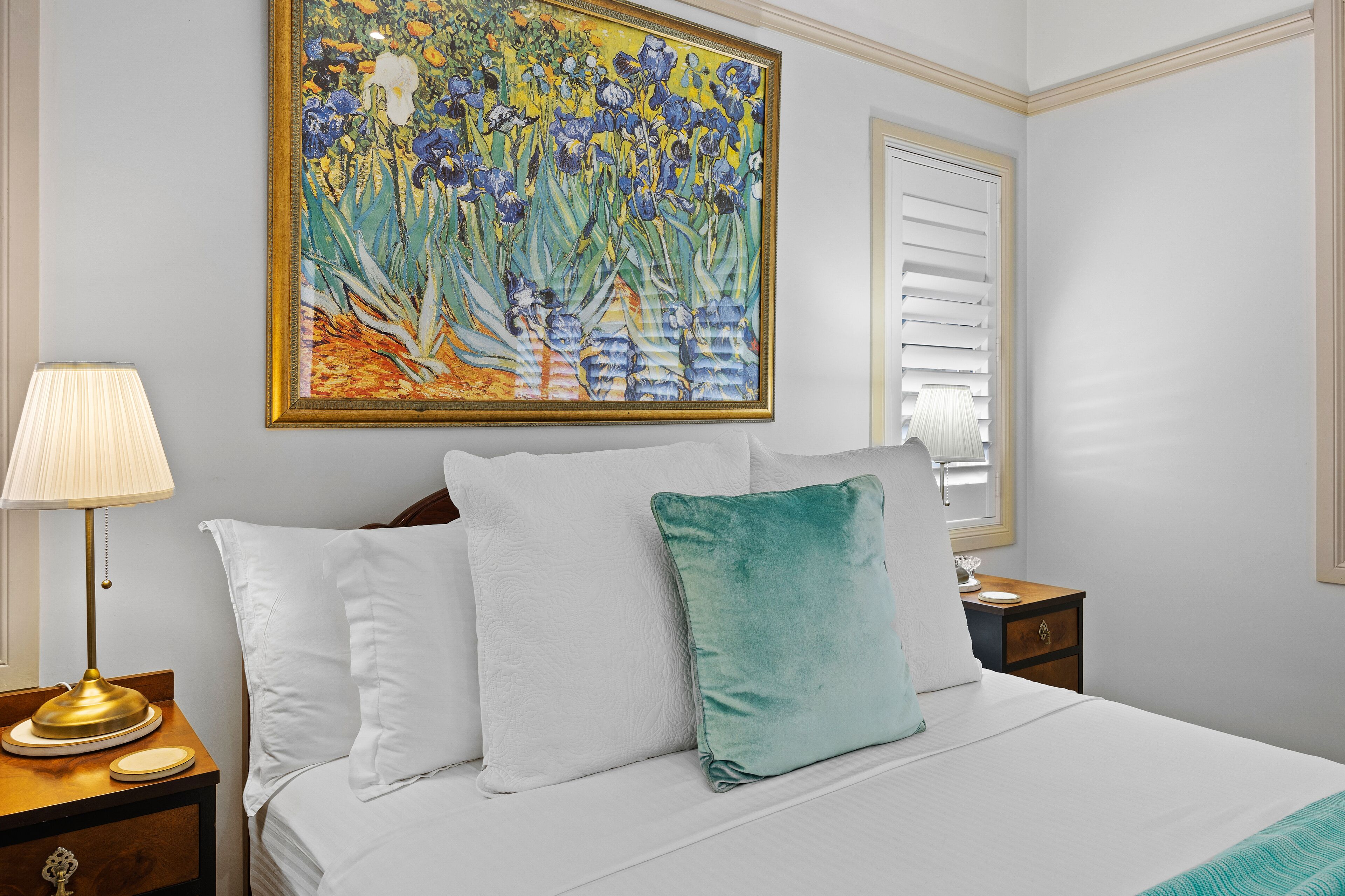 A neatly made bed with white and teal pillows, a bedside table with a lamp, and a large floral painting on the wall.