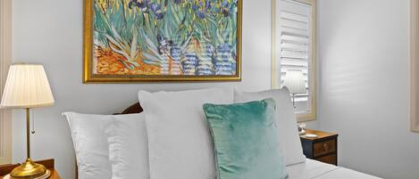 Una cama bien hecha con almohadas blancas y azul verdosas, una mesita de noche con una lámpara y un cuadro floral grande en la pared.