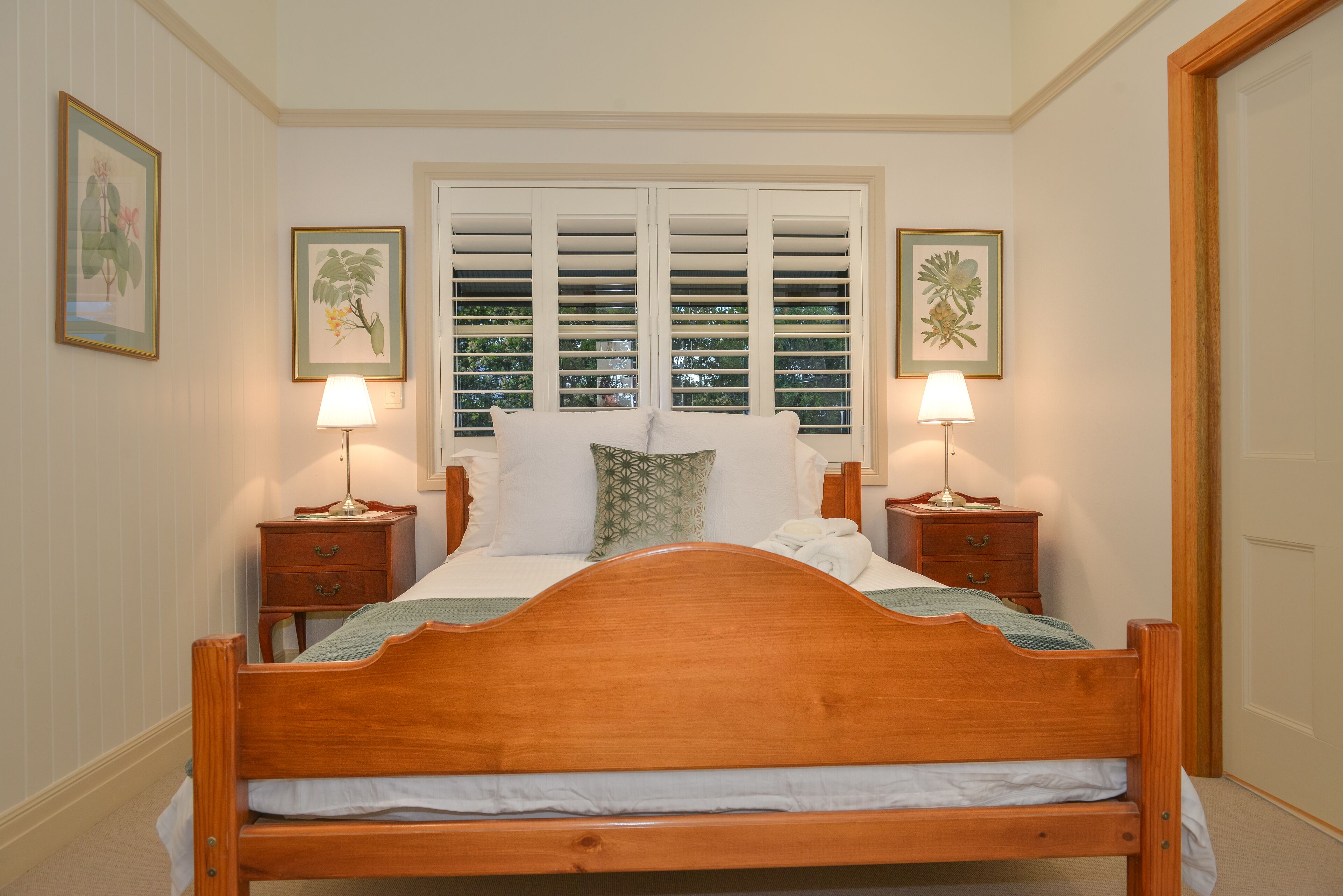 A bedroom with a wooden bed, two nightstands with lamps, a window with shutters, and framed botanical prints on the walls.