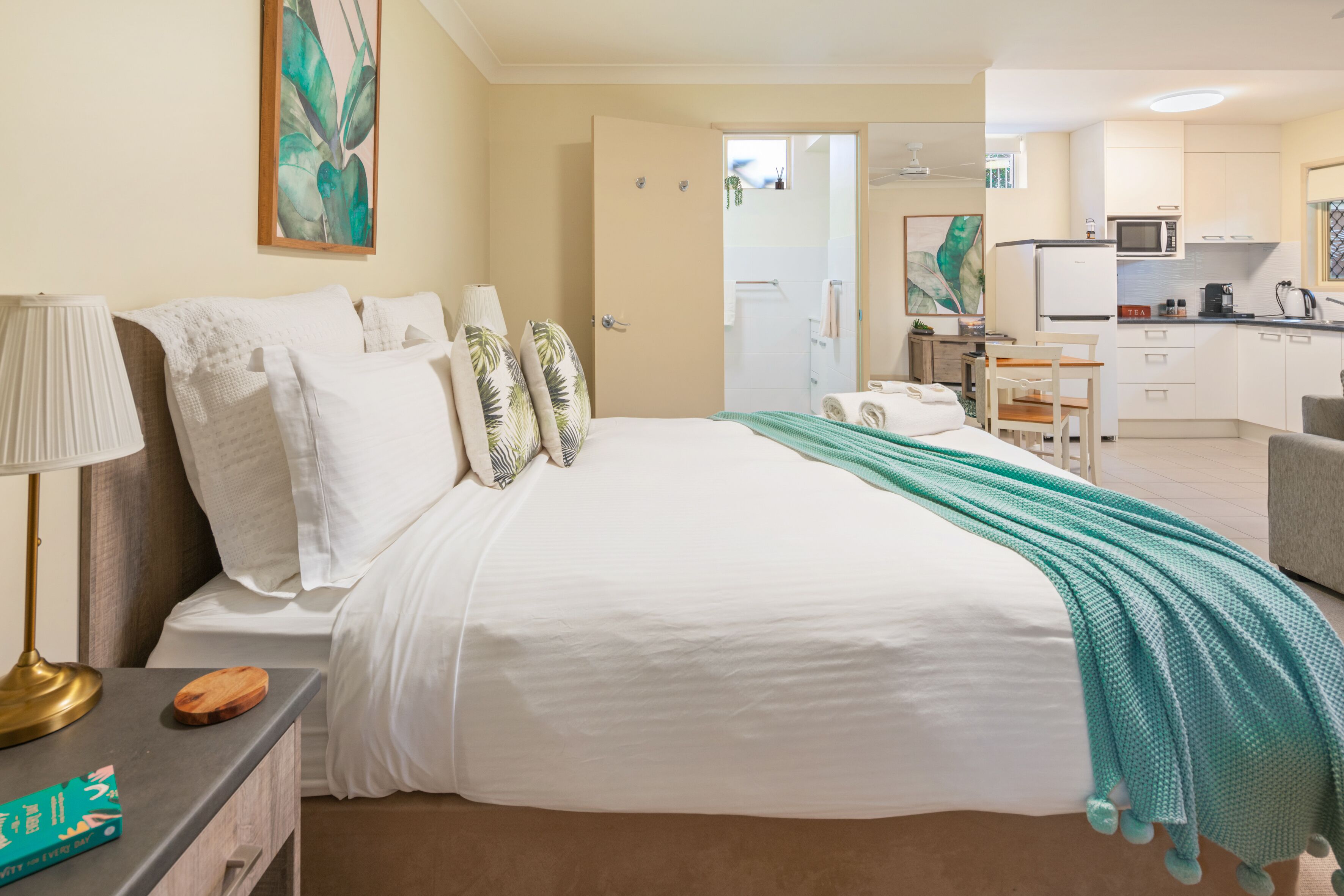 A hotel room with a bed, bedside table, lamp, and a view into the kitchen area.