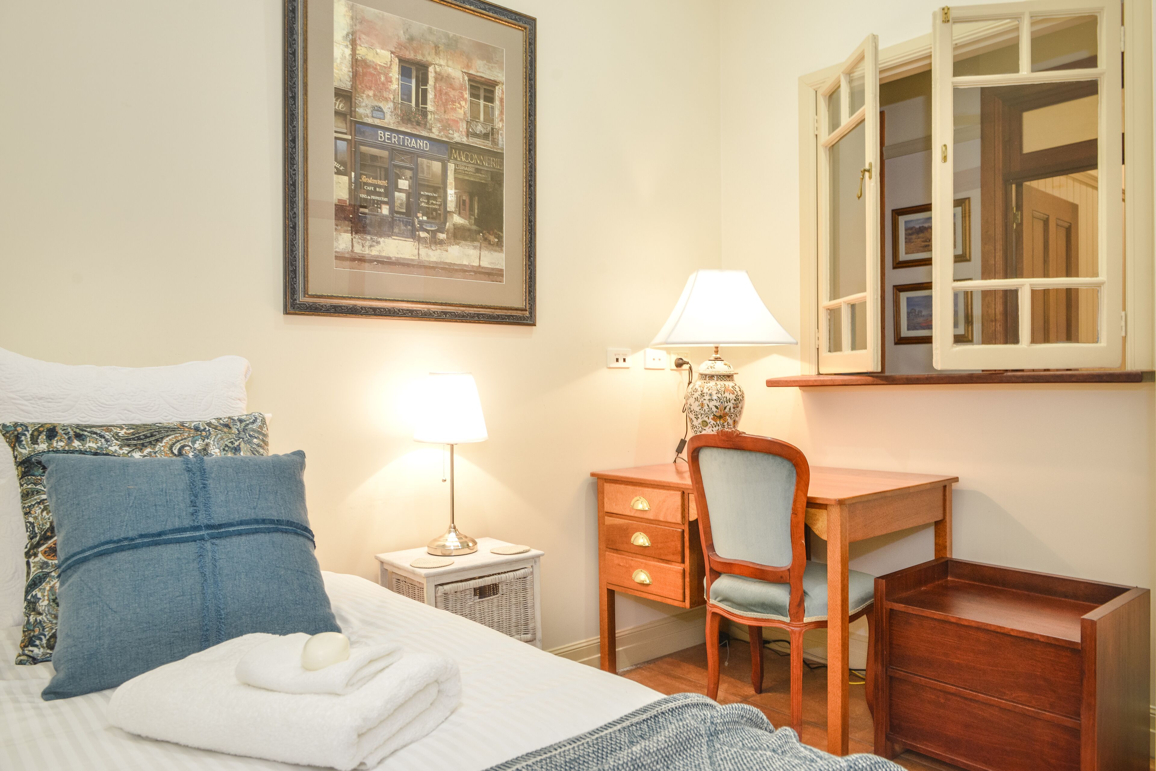 A neatly arranged hotel room with a bed, a desk, a chair, a lamp, and a framed picture on the wall.