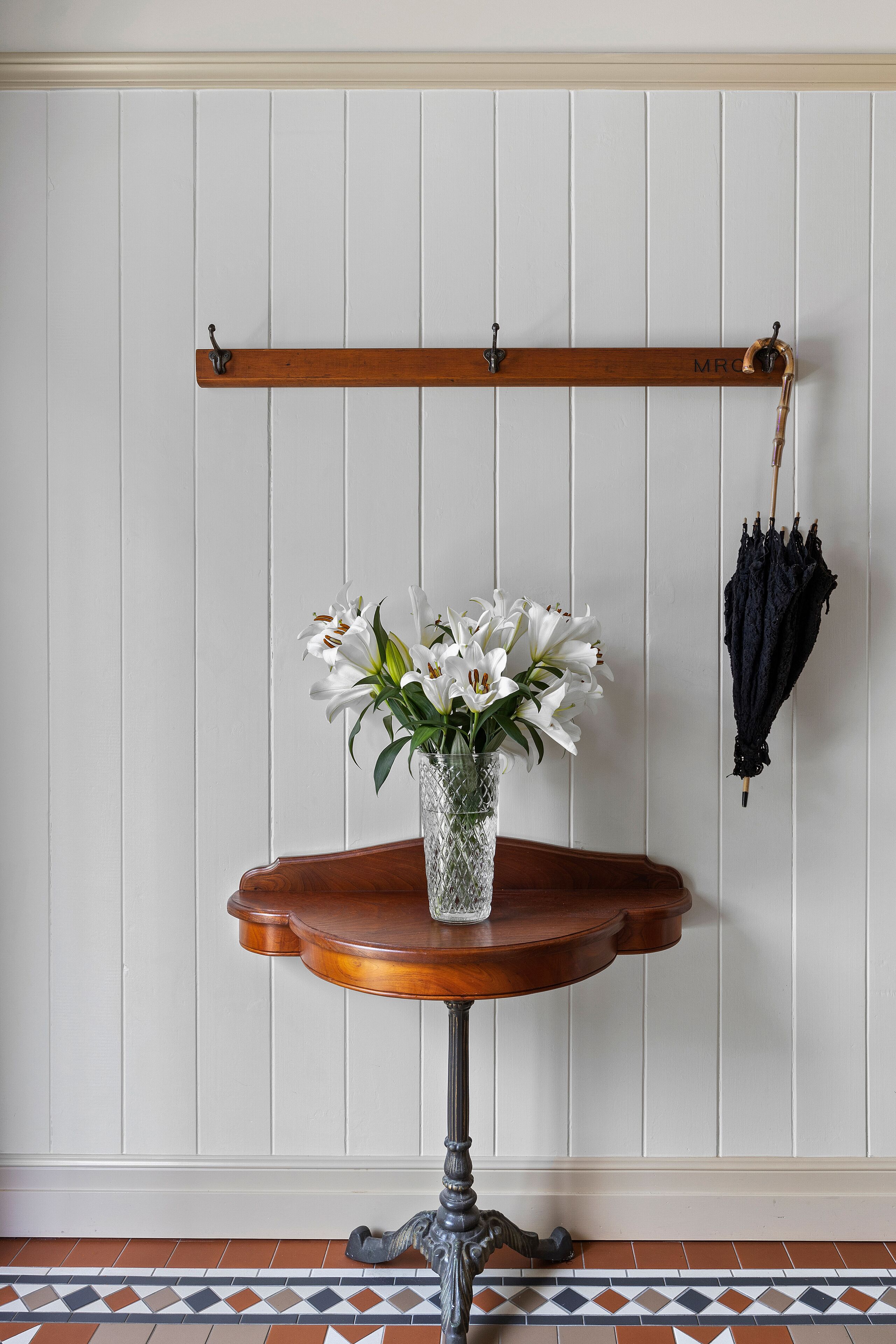 A wooden table with a glass vase of white flowers, a wooden coat rack with hooks, and a black umbrella hanging on the wall.
