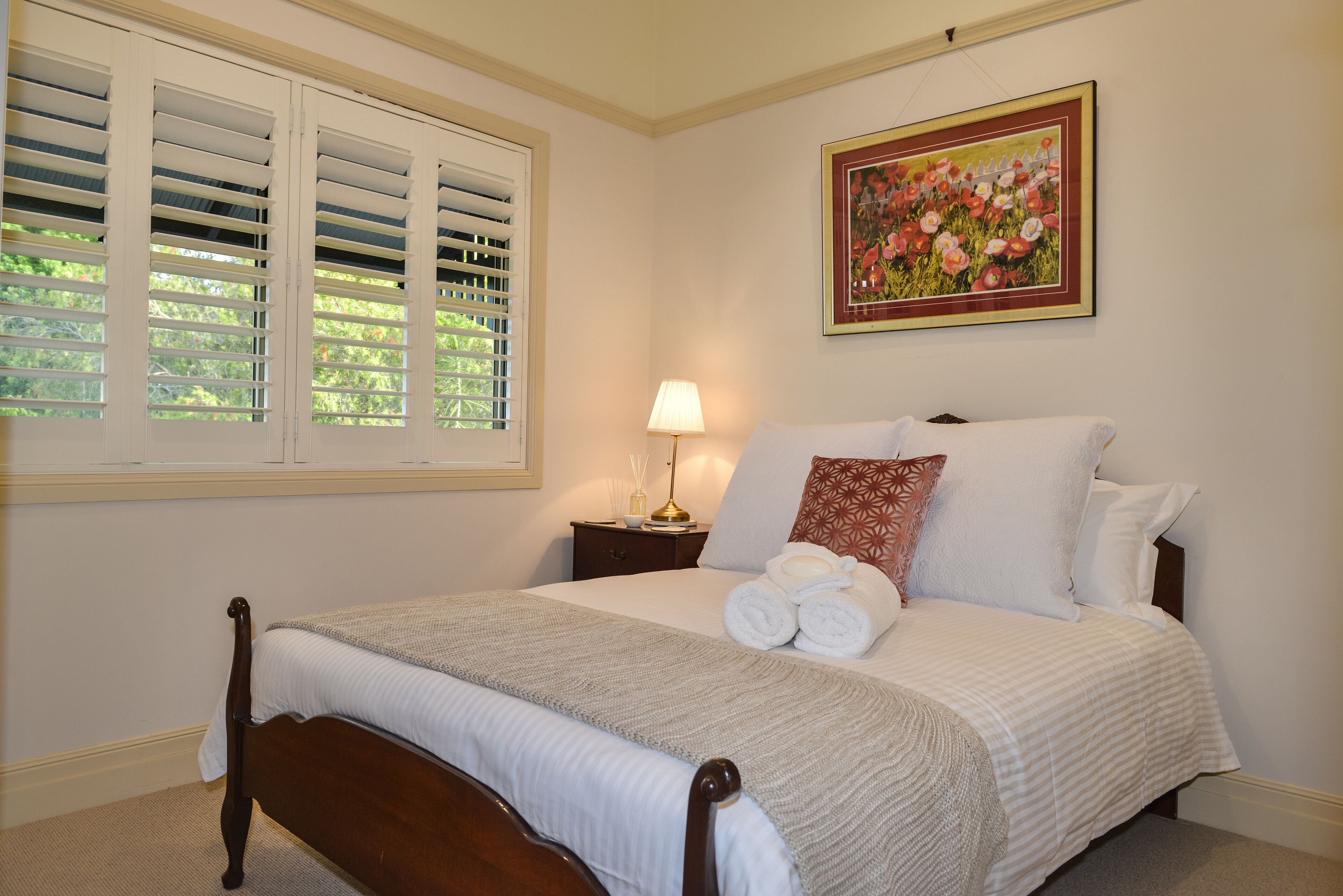 A neatly made bed with white linens and a gray blanket, a bedside lamp, and a framed floral painting on the wall.