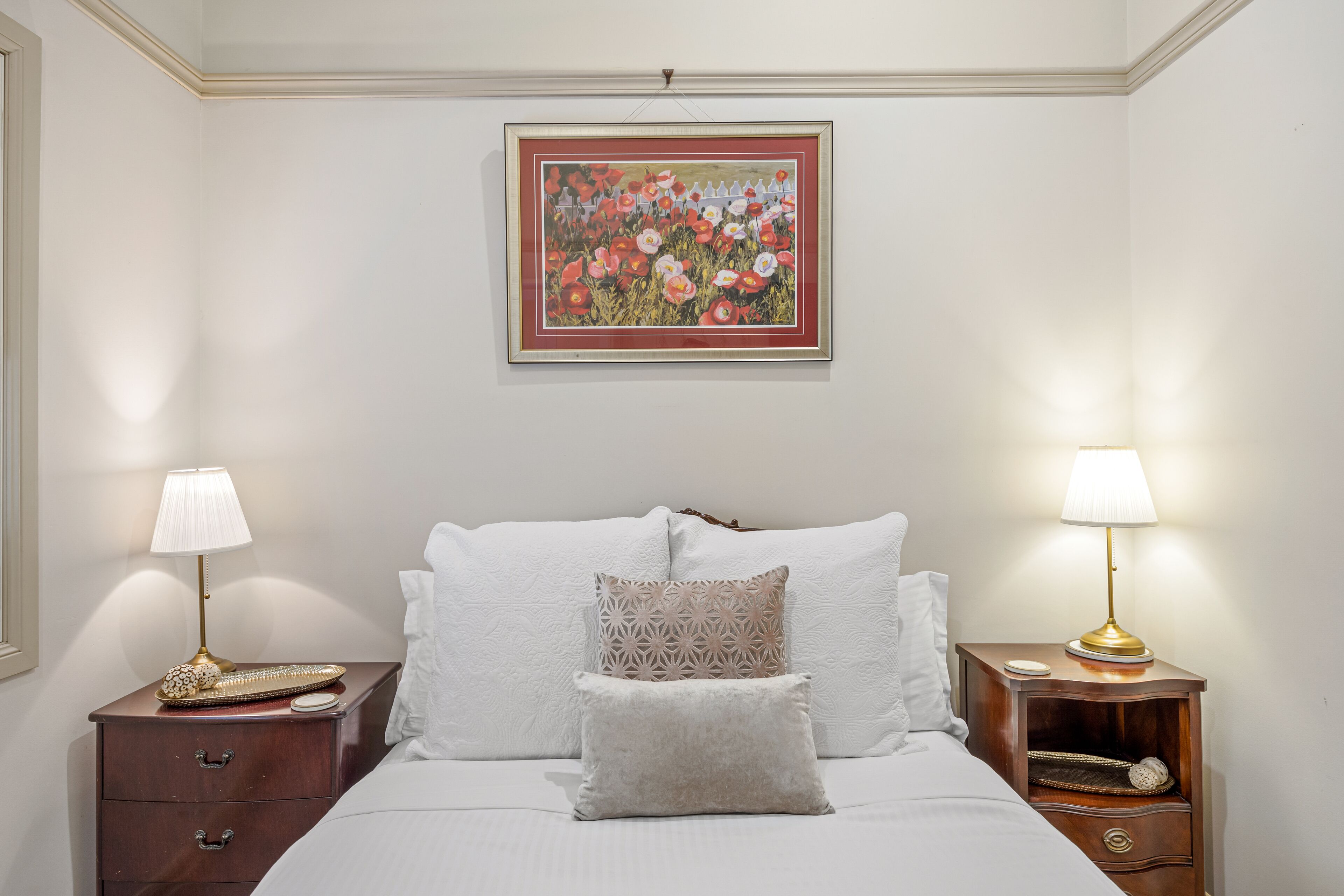 A neatly made bed with white linens, two bedside tables with lamps, and a framed floral painting on the wall.