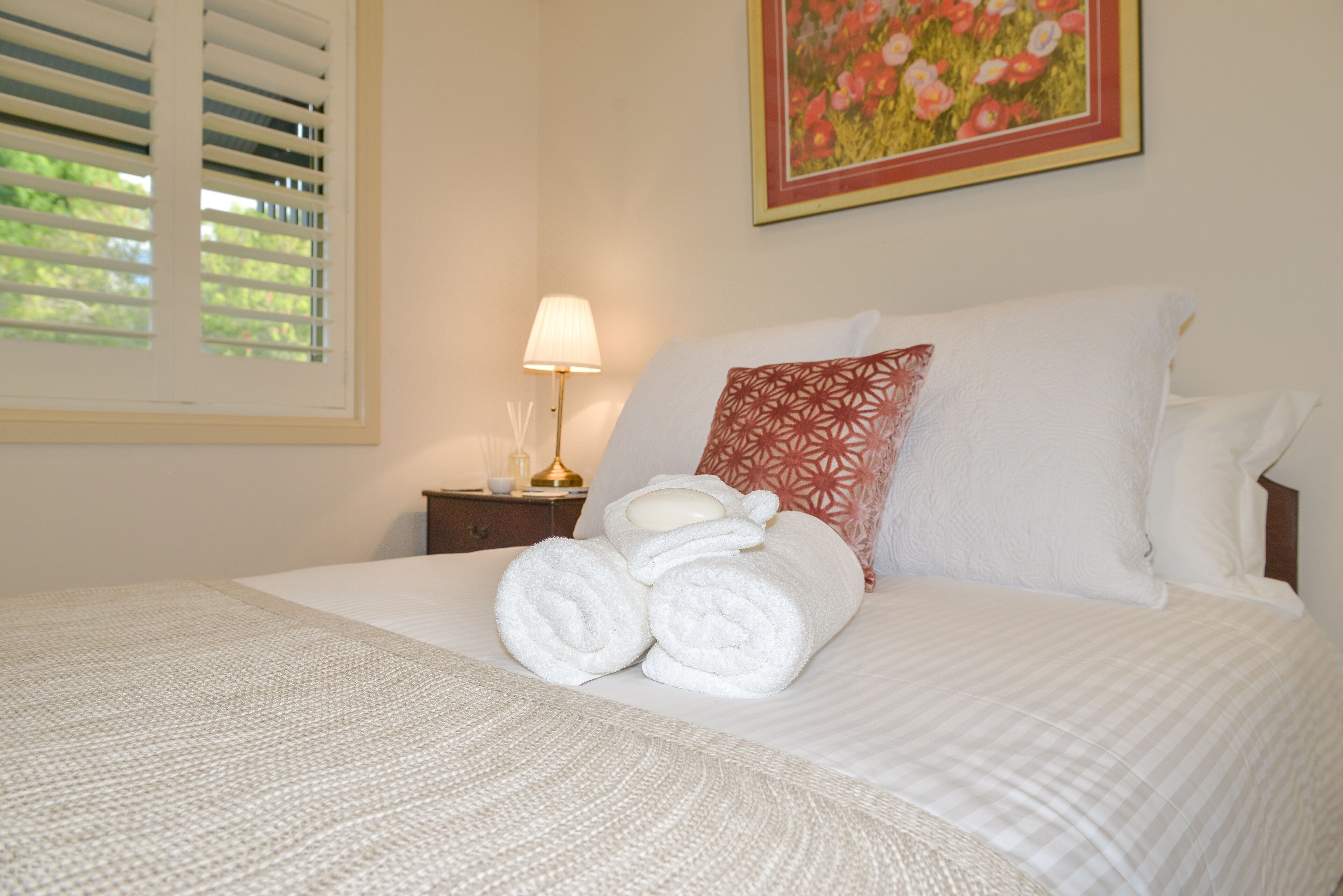 A neatly made bed with rolled towels, a bedside lamp, and a floral painting on the wall.