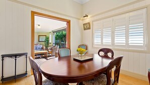 A dining room with a wooden table, floral chairs, a black console table, and a window with shutters. - The Guesthouse (Maleny)