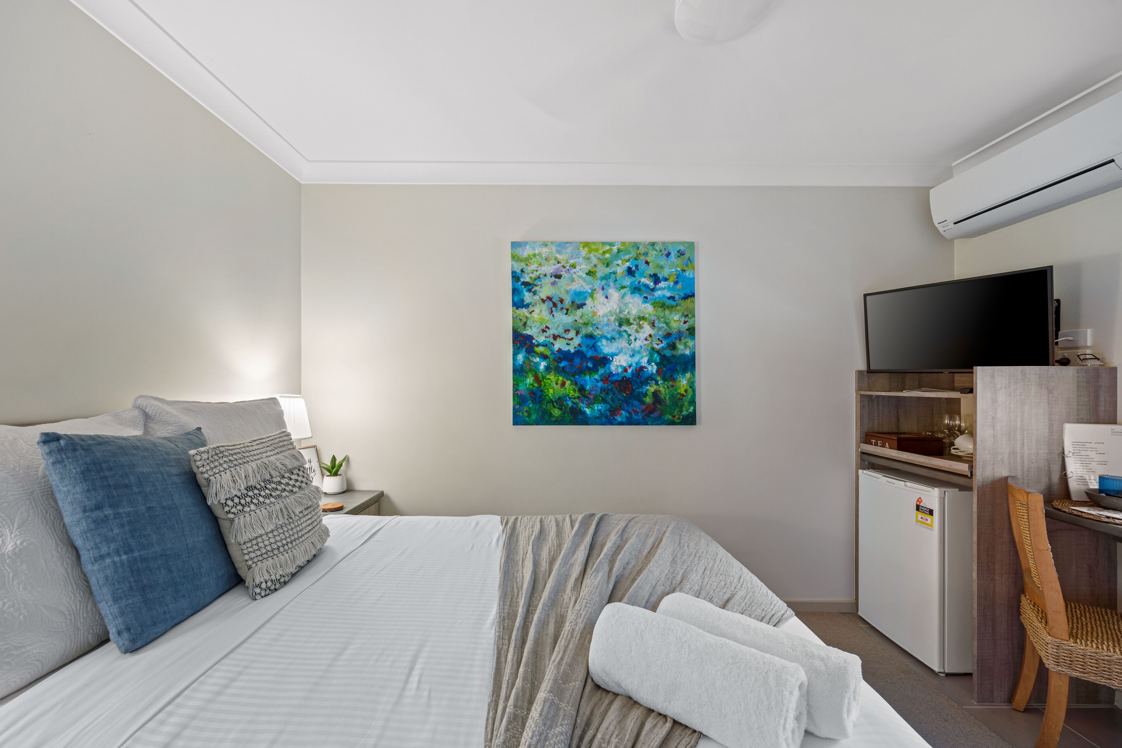 A modern hotel room with a bed, a desk, a television, and a painting on the wall.