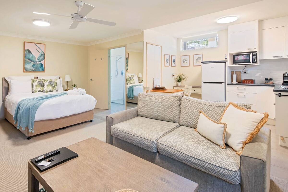 A compact hotel room with a bed, a sofa, a small table, a desk, and a kitchenette.