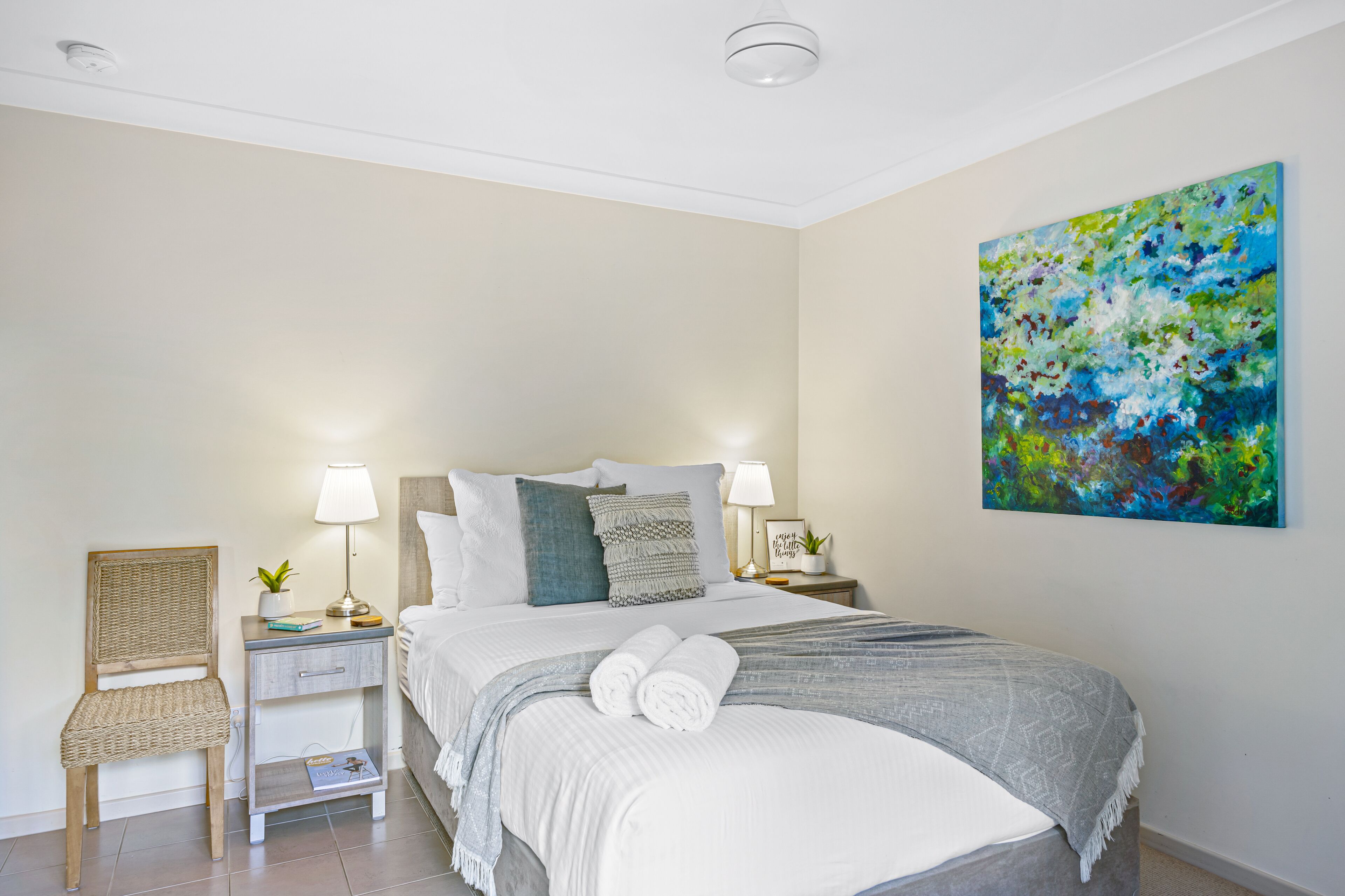A neatly arranged bedroom with a bed, bedside tables, a chair, and a painting on the wall.