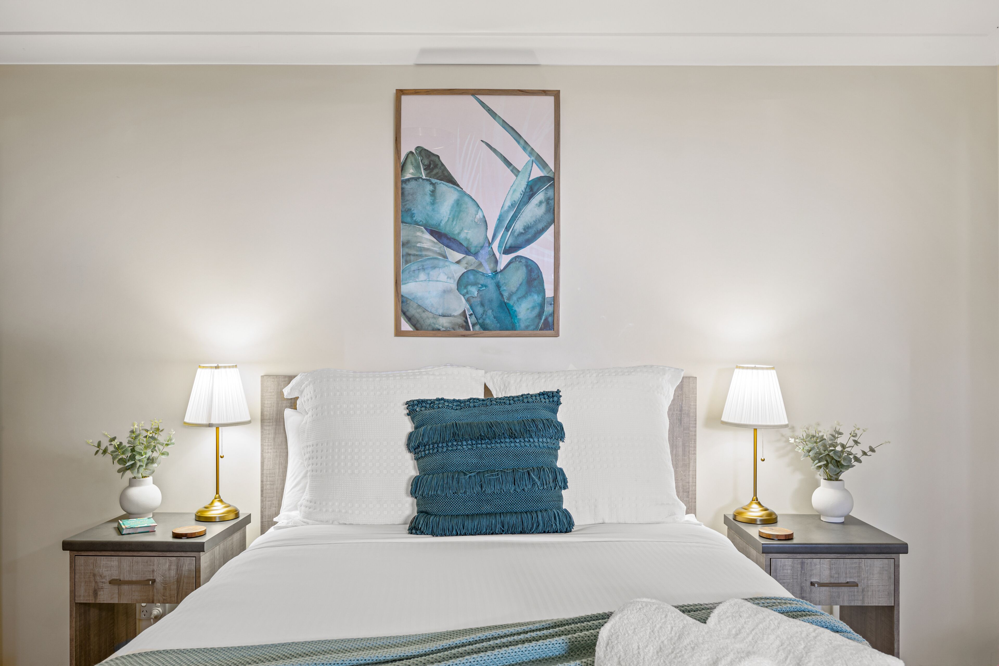 A neatly made bed with a blue pillow, two bedside lamps, and a framed artwork above.