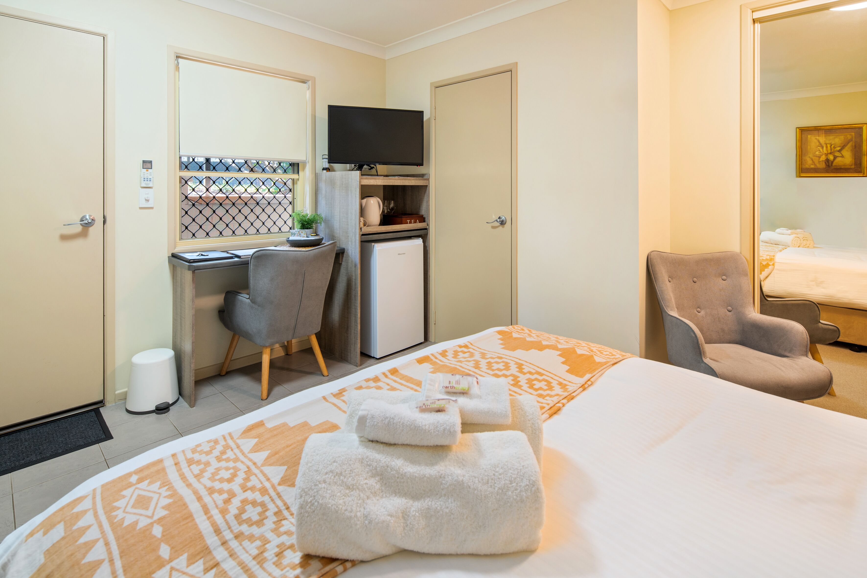 A hotel room with a bed, a desk with a television, a chair, a small refrigerator, and a bathroom visible through an open door.