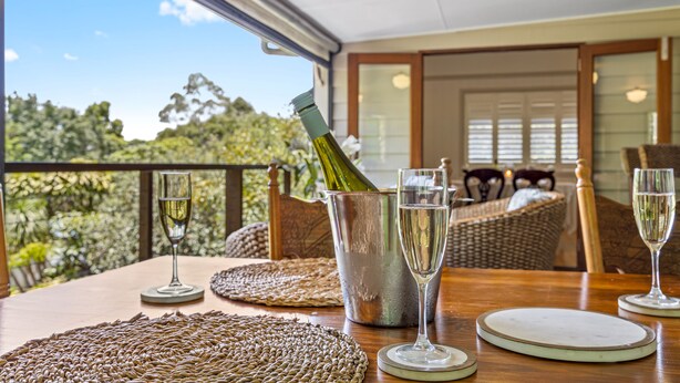 A dining table with a bottle of champagne, two glasses, and a metal bucket. The table is set with woven placemats and white plates. The room has wicker furniture and a view of trees through a glass door. - The Guesthouse (Maleny)