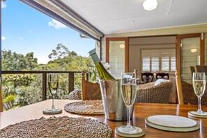 A dining table with a bottle of champagne, two glasses, and a metal bucket. The table is set with woven placemats and white plates. The room has wicker furniture and a view of trees through a glass door. - The Guesthouse (Maleny)